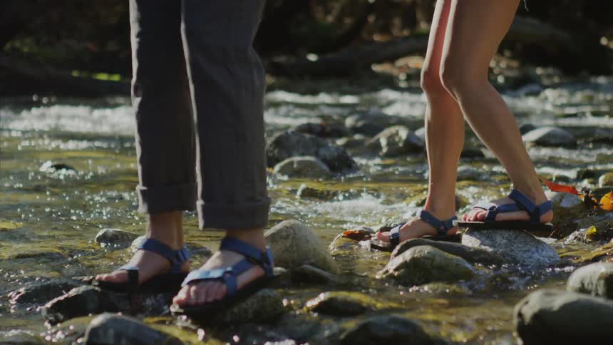 The feet of two people as they cross a river, balancing on slippery rocks