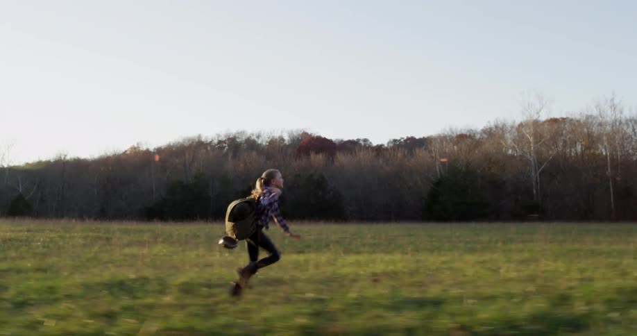 A young girl carrying a backpack is running across a field