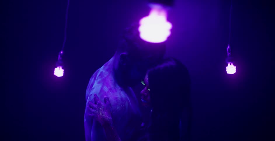 Lightbulbs swing around a man and woman covered in glowing body paint