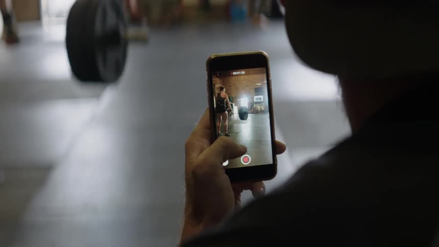 A man records a woman lifting weights in a gym