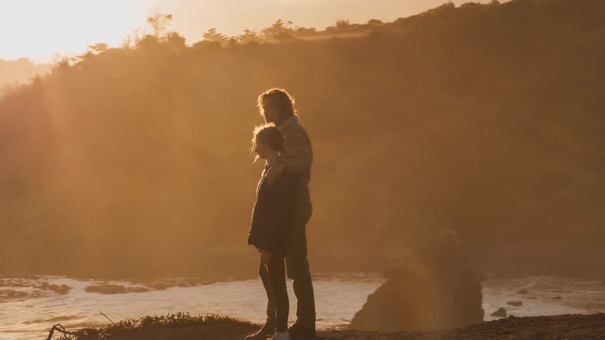 A man and girl stand on a cliff overlooking the ocean at sunset