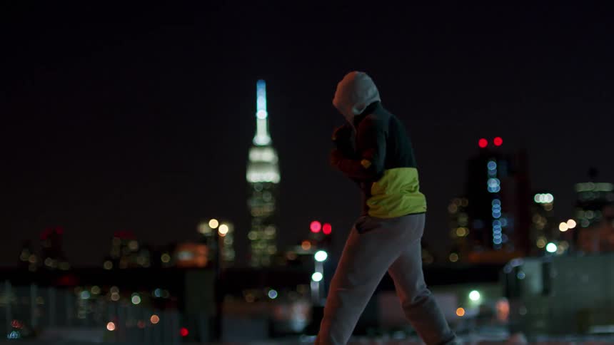 A man in a hoodie is shadow boxing at night on a roof with the city behind him