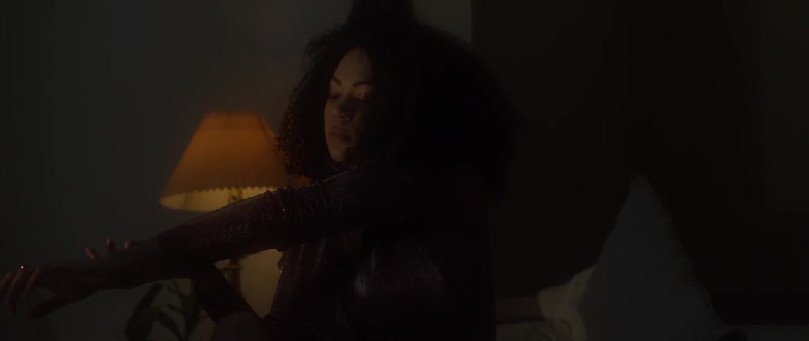 A woman sits on a bed in a dark bedroom and motions her arms around