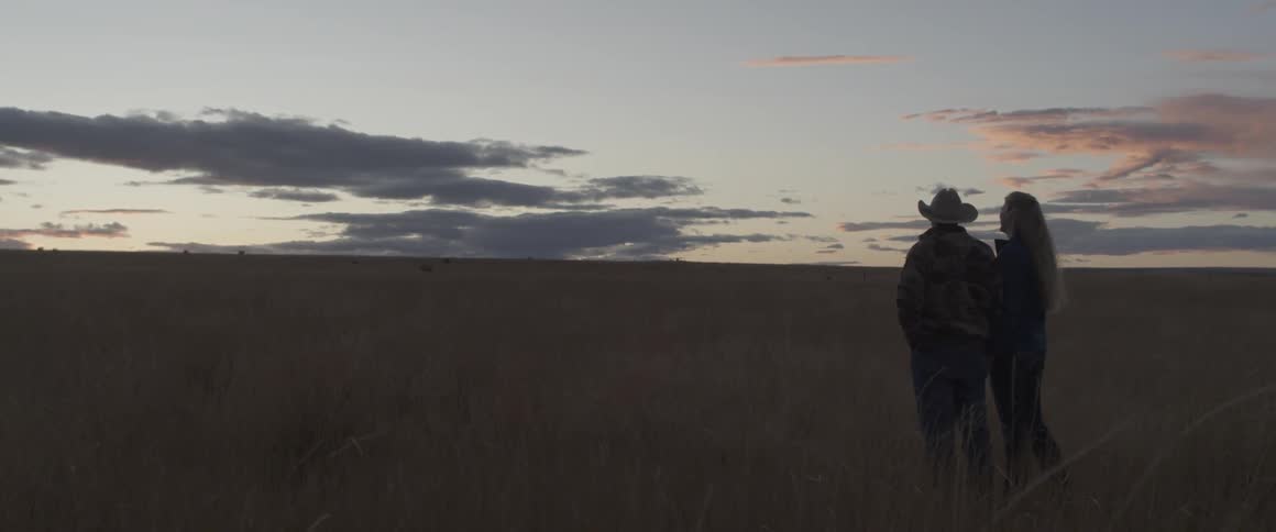 A cowboy and woman stand in a rural field at sunset