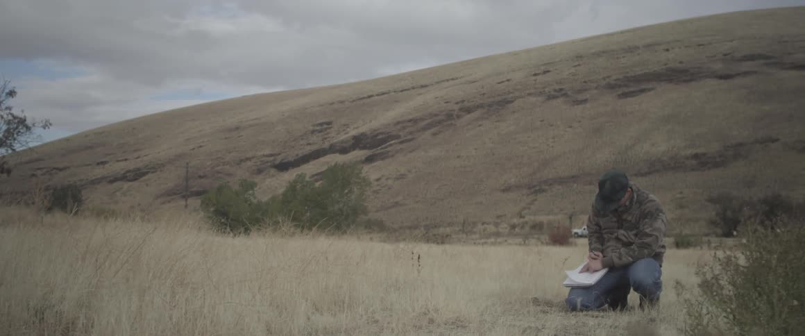 A man kneeling in the grass makes some notes