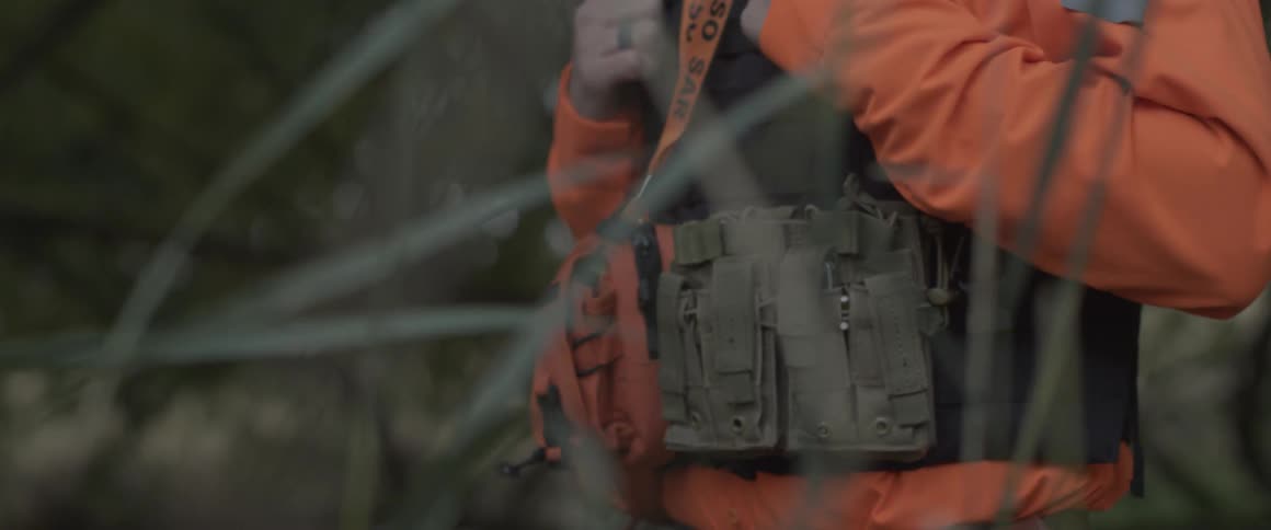 A man stands in a grassy area with a vest and utility belt