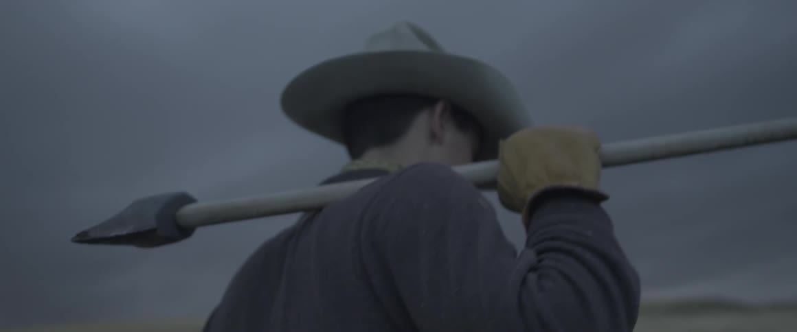 A cowboy carries an ax out in a field