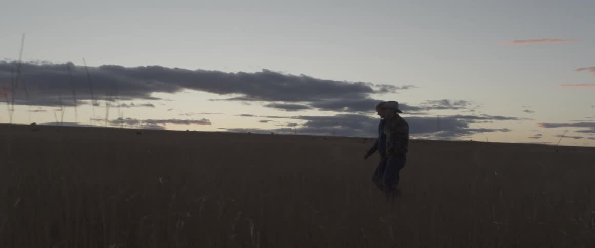 A cowboy and woman walk across a rural field at sunset