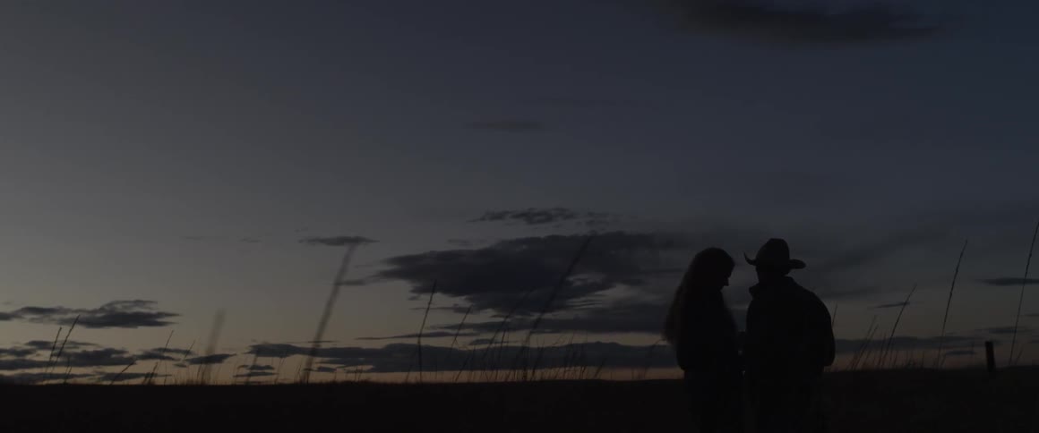 A cowboy and woman stand in a rural field at dusk