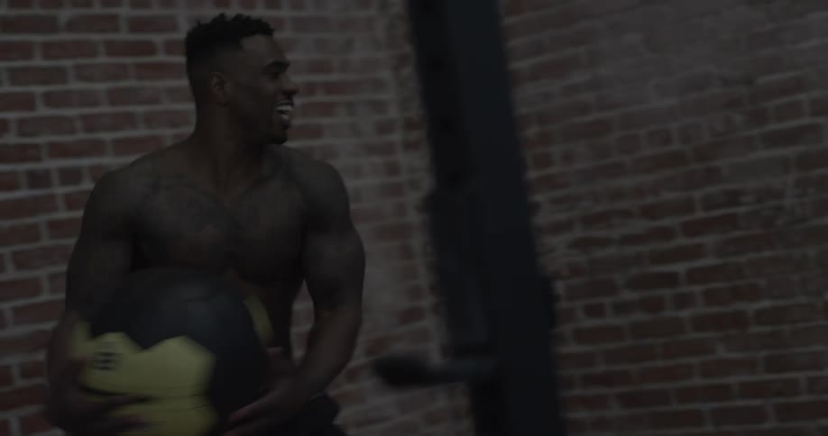 A shirtless man bounces an exercise ball off a brick wall and catches it