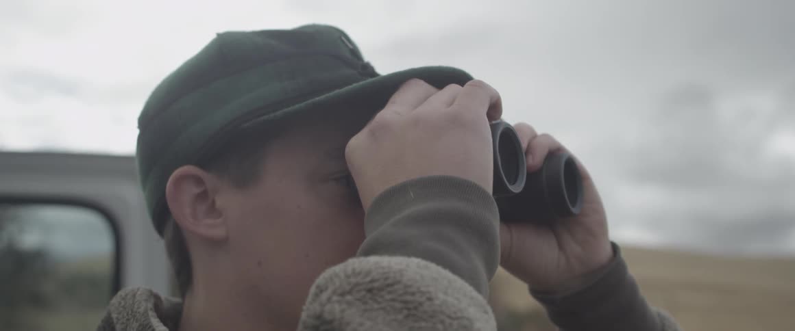 A man wearing a cap stands outside looking through binoculars