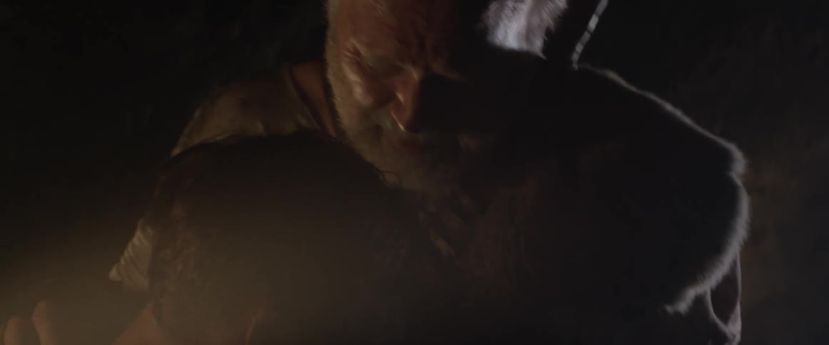 An elderly man puts a necklace on another man in a dark cave