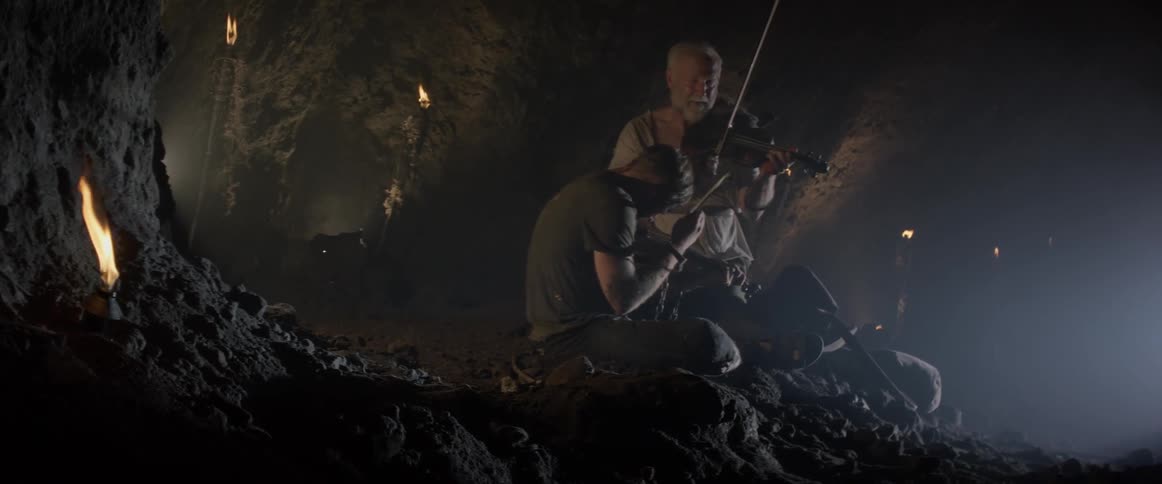 An old man encourages a bloodied man to play a violin as they sit in a cave