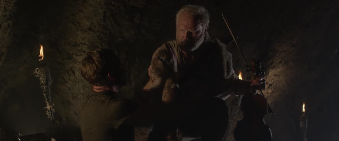 An elderly man tends to a hurt young man in a cave