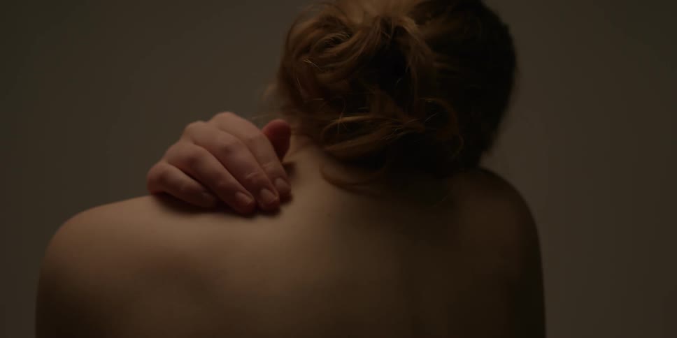 A shirtless woman wraps her arms around her shoulders and touches her head