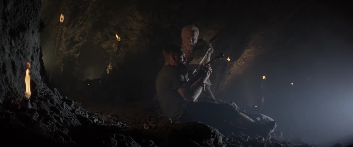 Two men play violins in a cave lit by torches