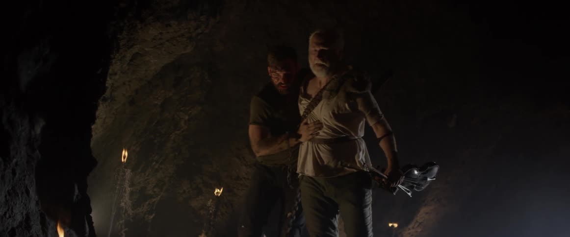 A bloodied man leans on an older man as they walk out of a cave