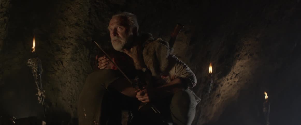 An elderly man crouches and hugs an injured man in a dark cave