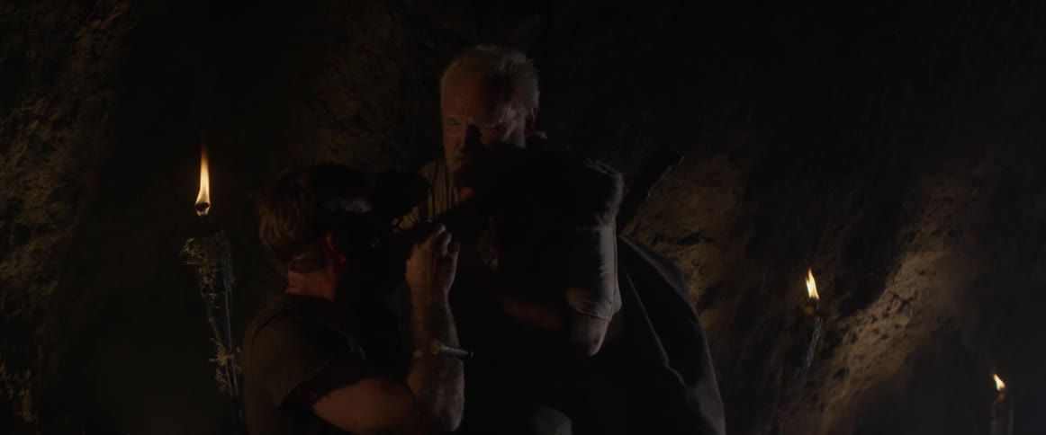 An older man puts a necklace on a bloodied man in a cave