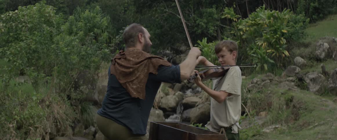 A man and boy play violin next to a stream in the woods