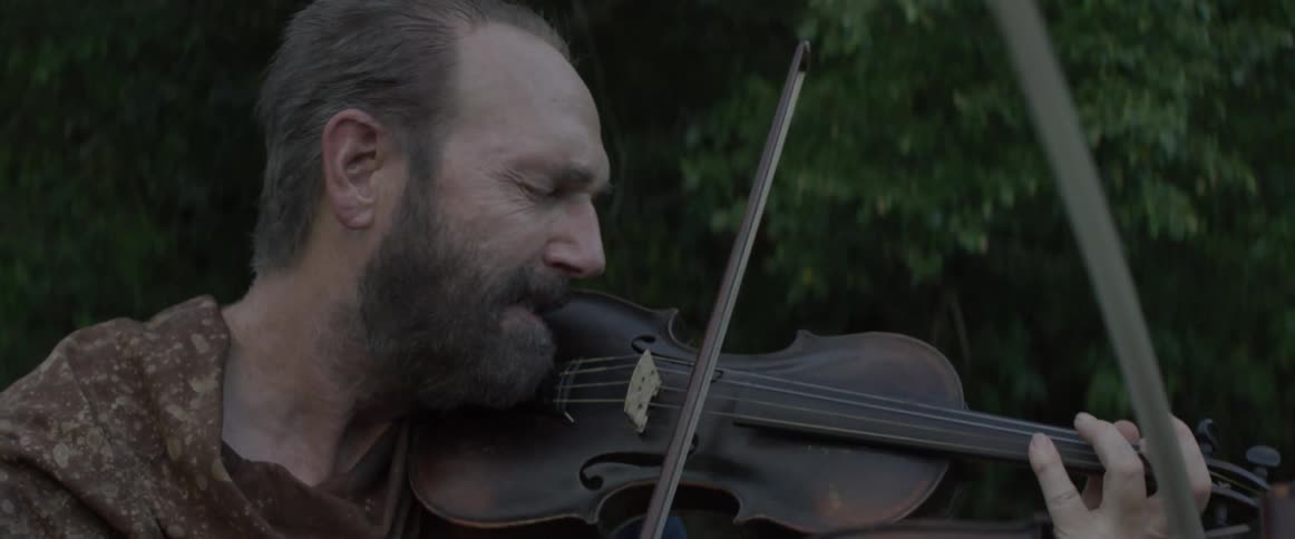 A man plays violin with another person outside near trees