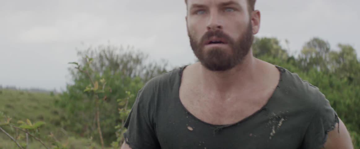 A man in a tattered shirt walks near shrubs and trees