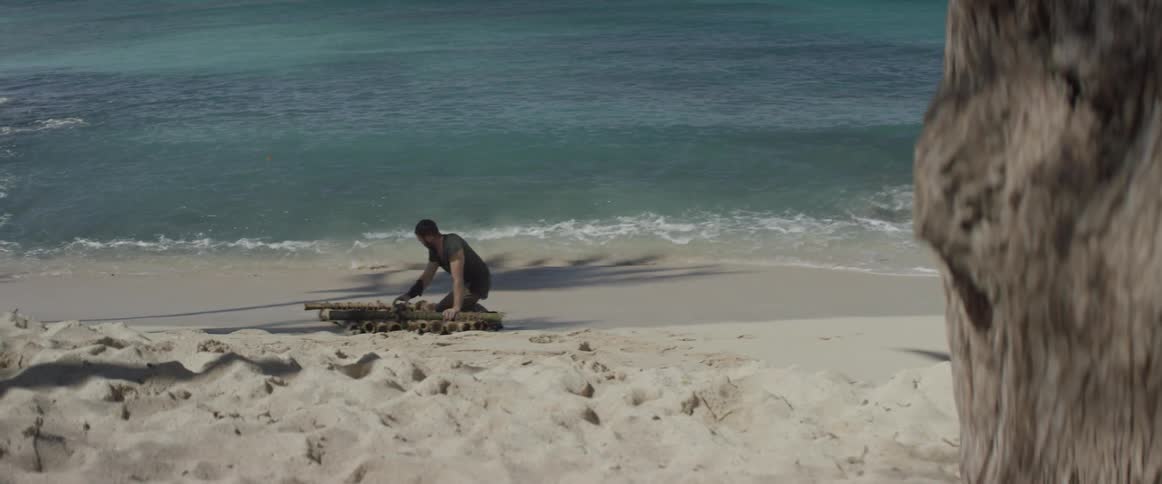 A castaway kneels on a wood raft on a beach