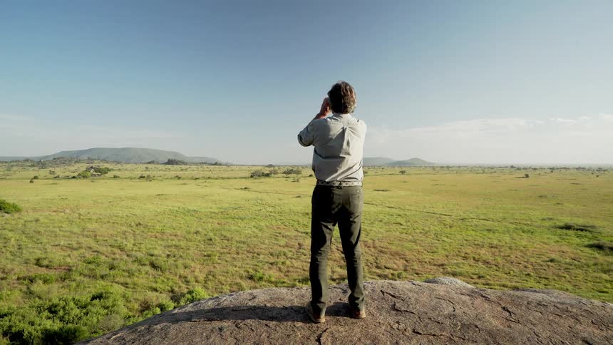 A man walks atop a hill and looks out with binoculars