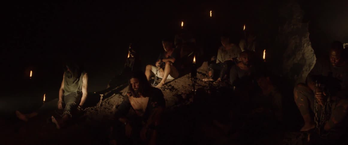 A group of injured prisoners sit in a dark cave