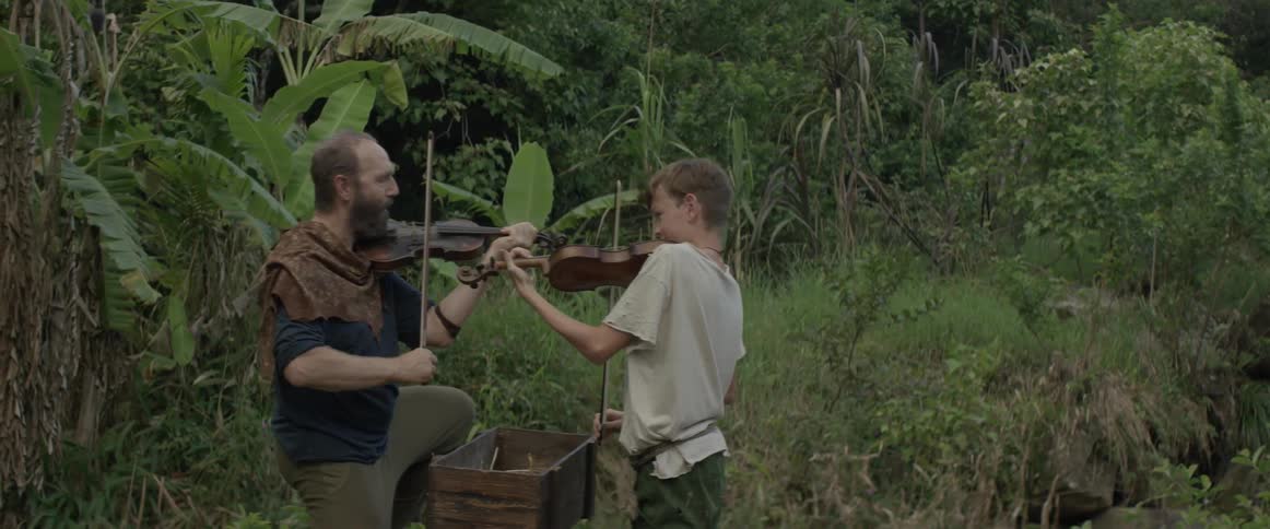 A man and a young boy kneel on the ground in a forest area and play the violin