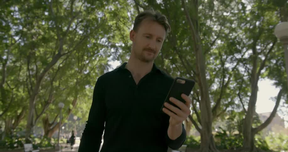 A man walks outside under trees looking at a cell phone