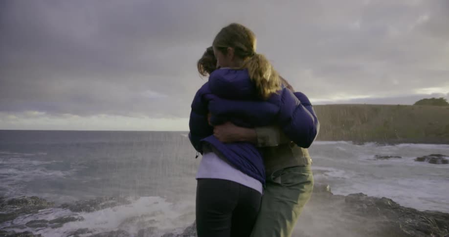 A man and girl jump backward as an ocean wave crashes ashore