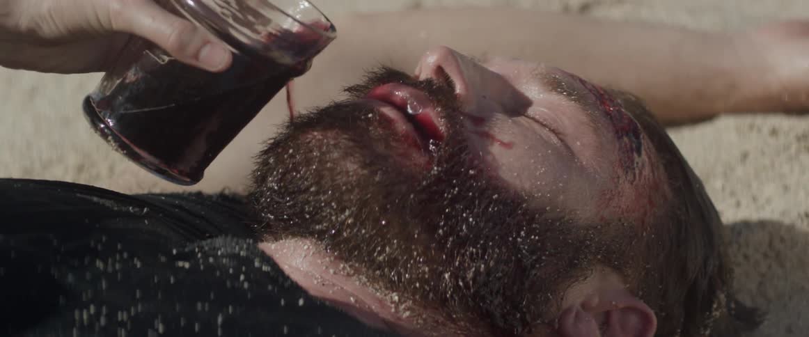 Man lays on the sand as someone pours a drink into his mouth
