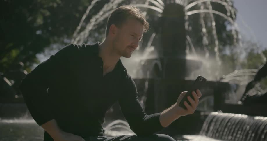 A man sits by an outdoor water fountain and types on his cell