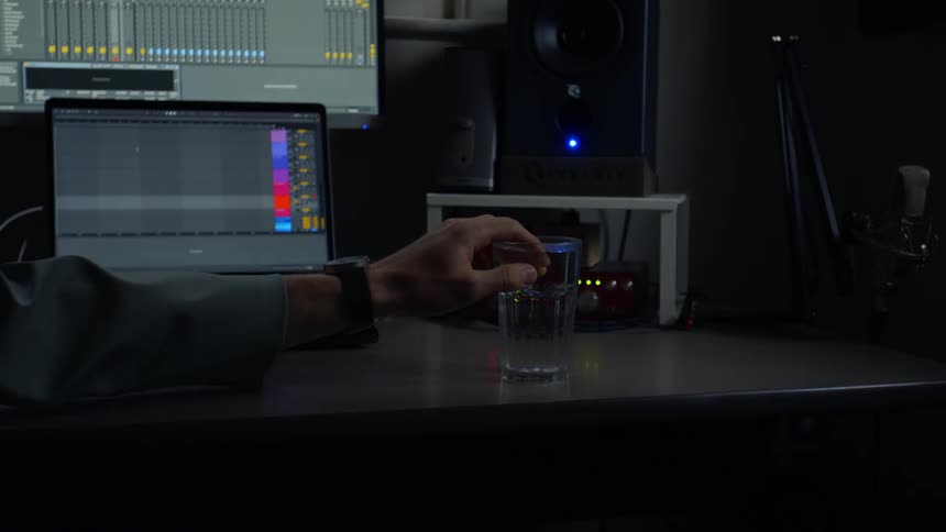 Person's hand on a desk reaches towards a glass with computer equipment in the background