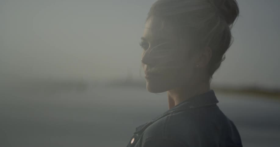 A woman with her hair up is smirking at the camera standing on the beach