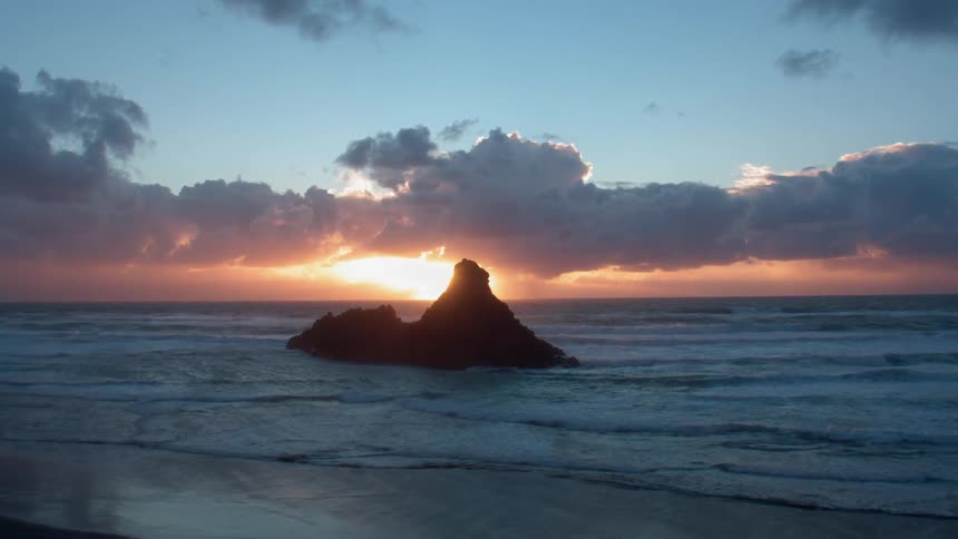 A drone filming a rock sticking out of the ocean with the sunset in the background