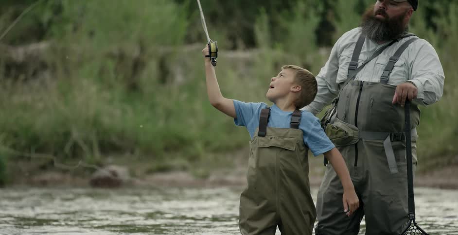 A man helps a boy cast a fishing line in a river during the day