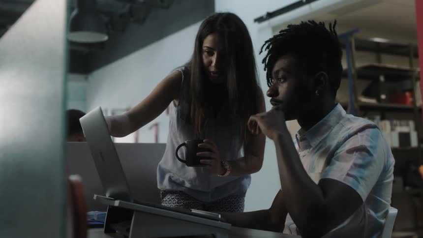 A woman walks over and talks to a man working at a computer in an office