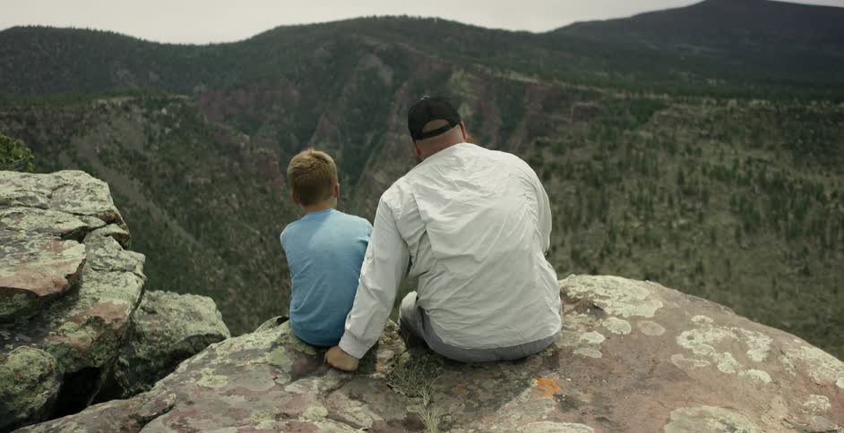 A man and a young boy sit at the top of a mountain