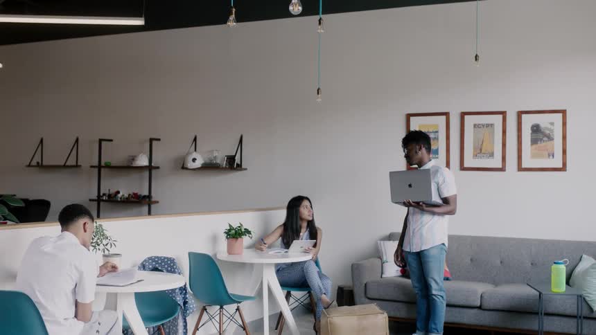 Man holding a laptop talks to a woman sitting at a table in a cafe