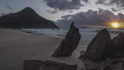 Timelapse of a beach with rocks, a mountain in the background, and rolling clouds during sunrise
