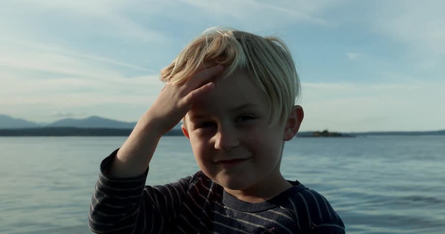 Child brushes hair off his head standing next to a body of water