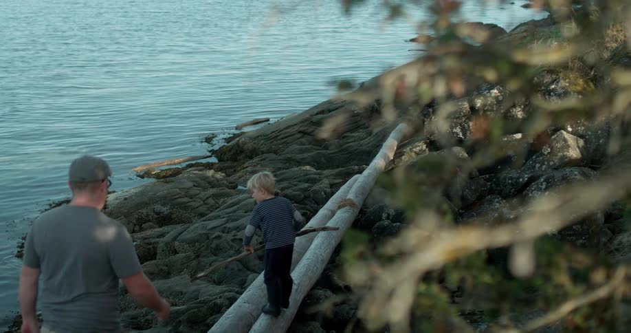 Child carries a stick and walks on logs near water with adult following
