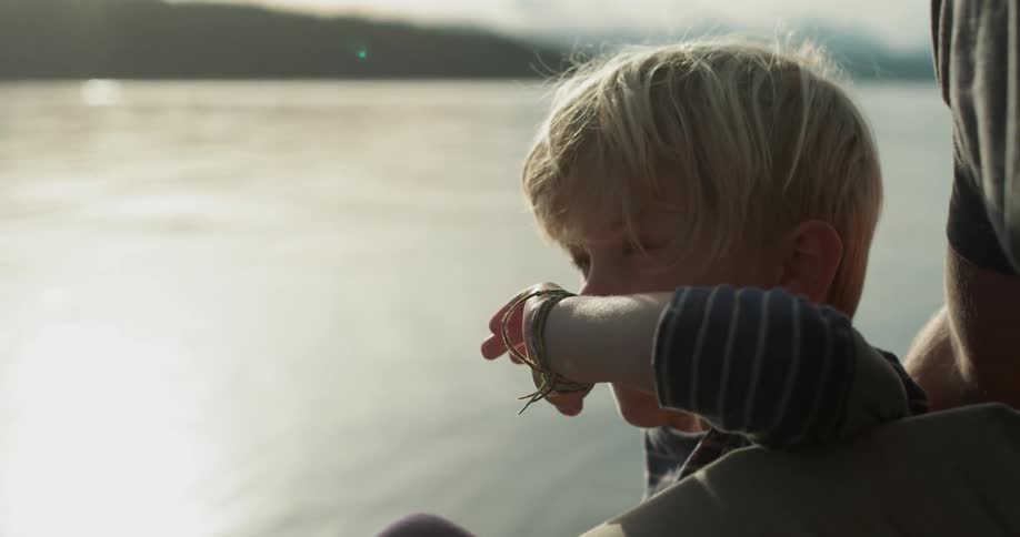 A boy sits near the water leaning against his dad