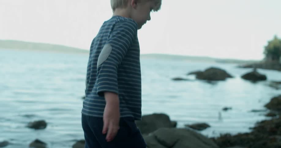 A boy wearing boots walks on the rocks along the water