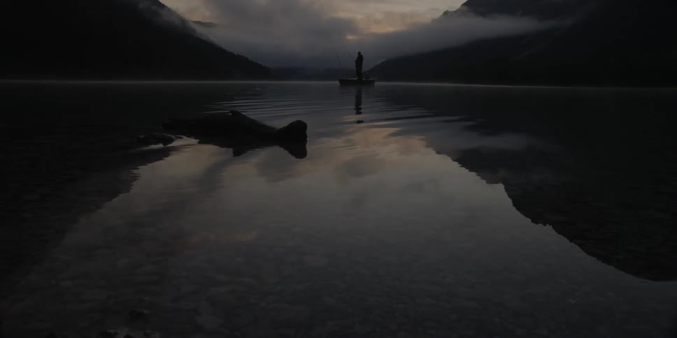 A man stands in a boat fishing at dusk or dawn
