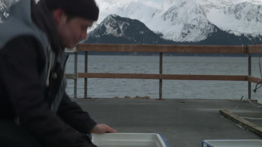 A man squats and investigates the contents of a container, then walks over to a wooden railing