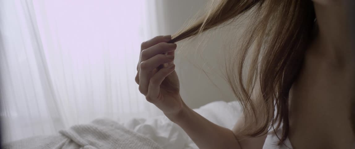 A woman sits in bed feeling her hair with her hand