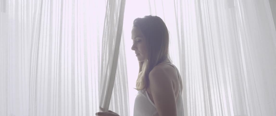 A woman stands next to a window holding onto curtains